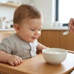 Un bébé souriant assis dans une chaise haute en bois devant un bol de yaourt pendant qu'un parent lui donne à manger.